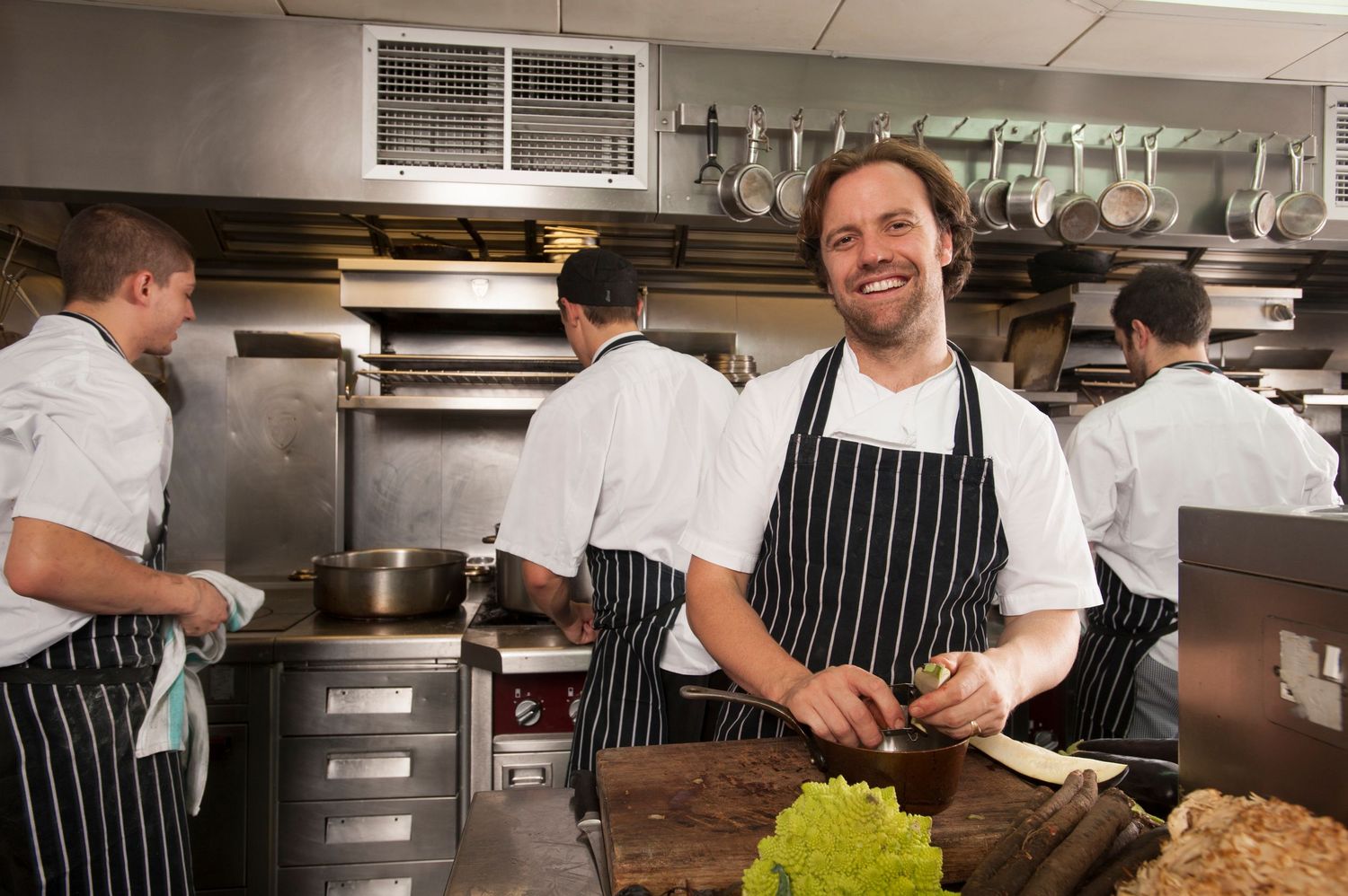 Smiling chef in striped apron prepares food in a busy professional restaurant kitchen with colleagues cooking in background.