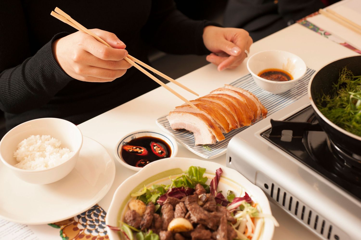 Person using chopsticks to eat sushi and Asian dishes with rice, soy sauce, and greens at a restaurant table.
