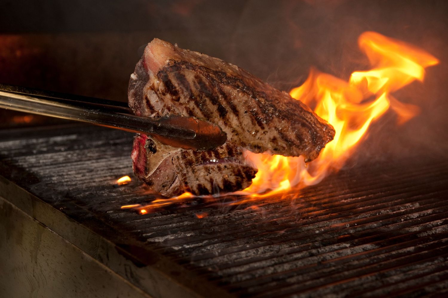 Juicy grilled steaks sizzling over open flames on a barbecue grill, held with tongs for perfect char.