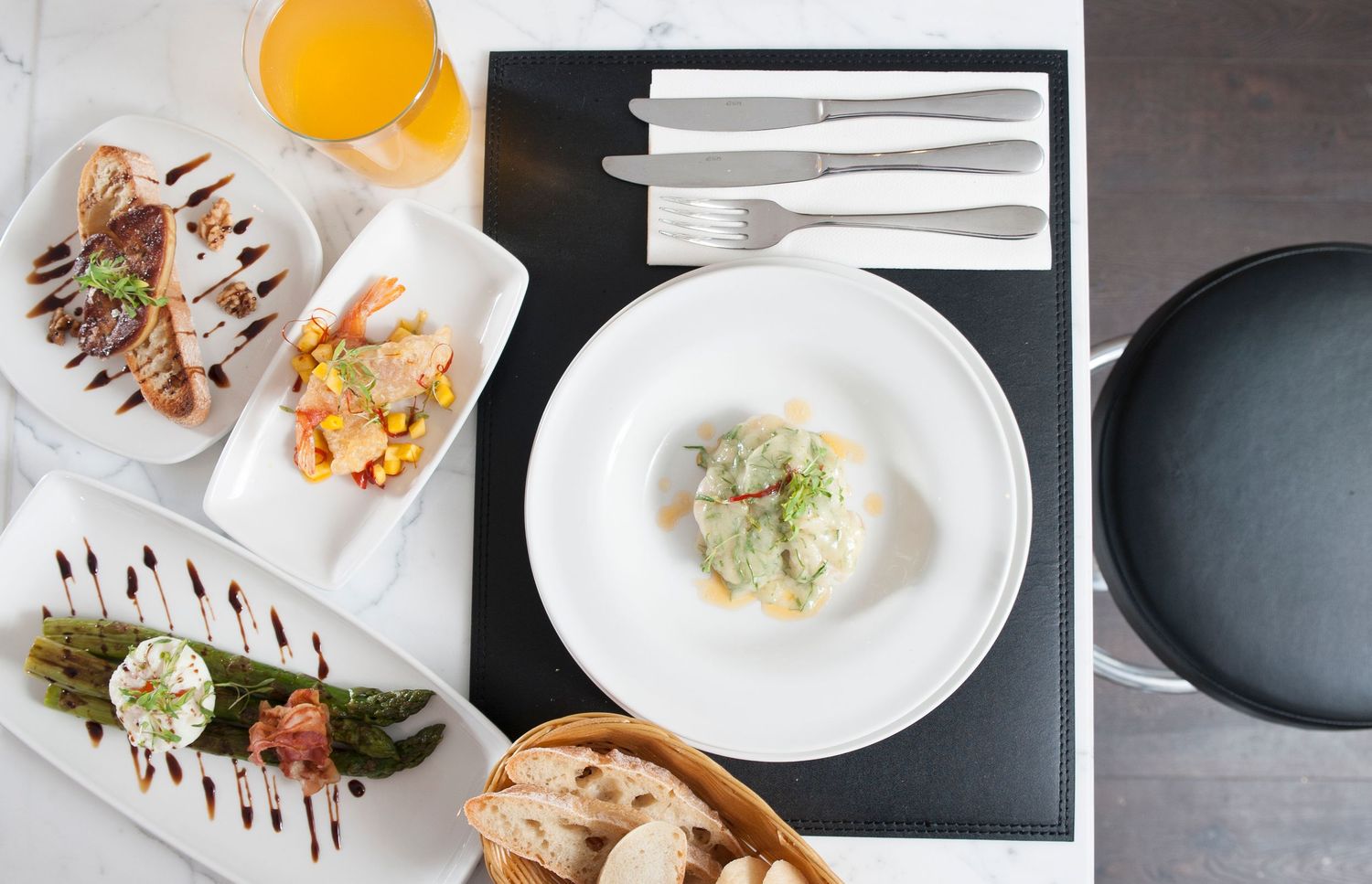 Elegant restaurant table setting with gourmet dishes, fresh bread, orange juice, and fine cutlery on a marble surface.