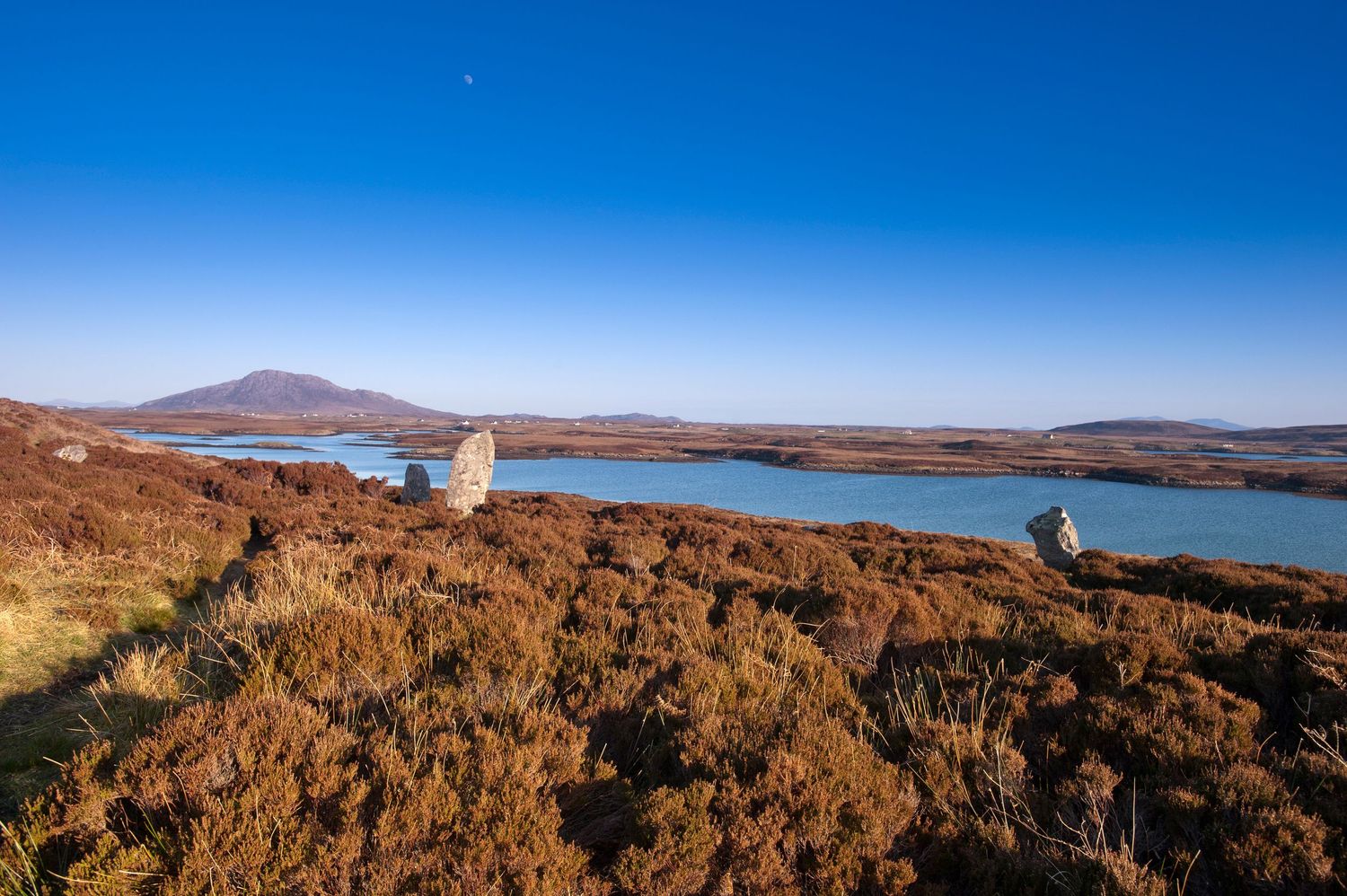 Panoramic view of a Scottish loch with standing stones amid heather moorland under a vivid blue sky.