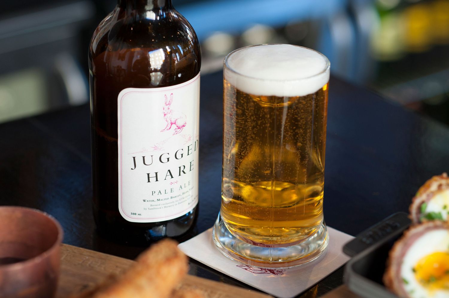 Juggling Hare Pale Ale bottle beside a full pint glass on a coaster, with a Scotch egg in the background.