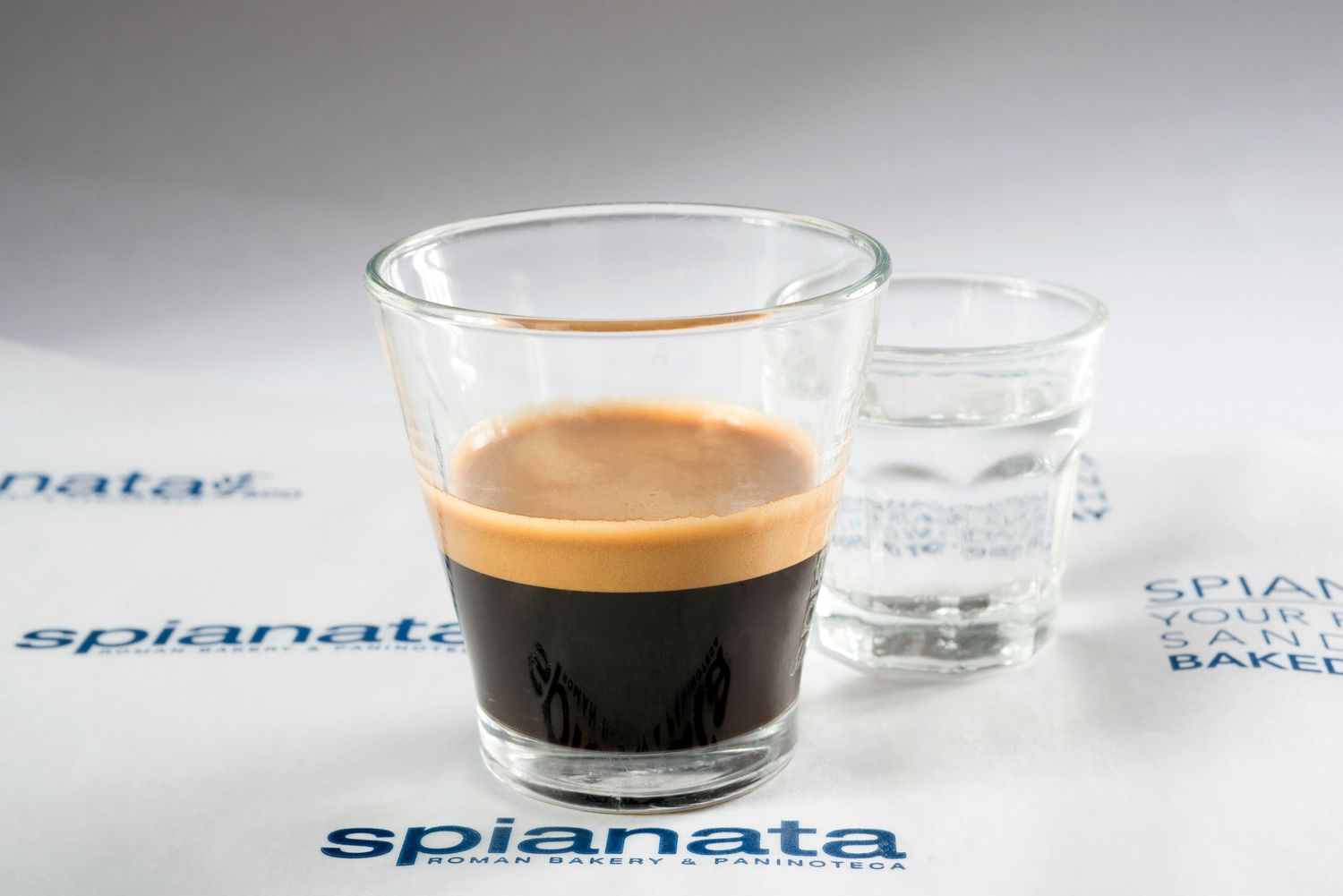 A rich espresso in a glass cup beside water on a Spianata branded tablecloth.