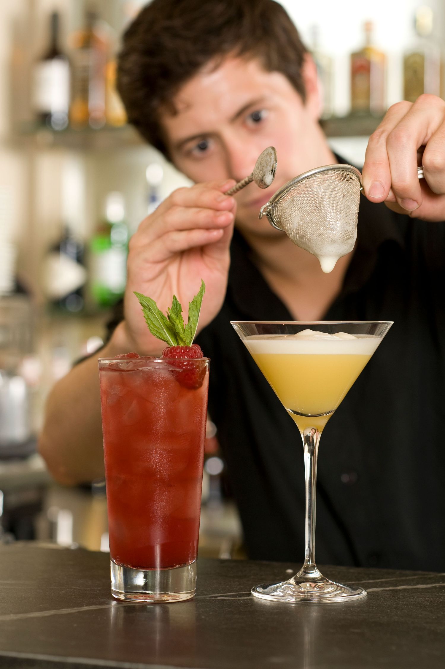 Bartender straining a yellow cocktail into a martini glass beside a red berry drink garnished with mint.