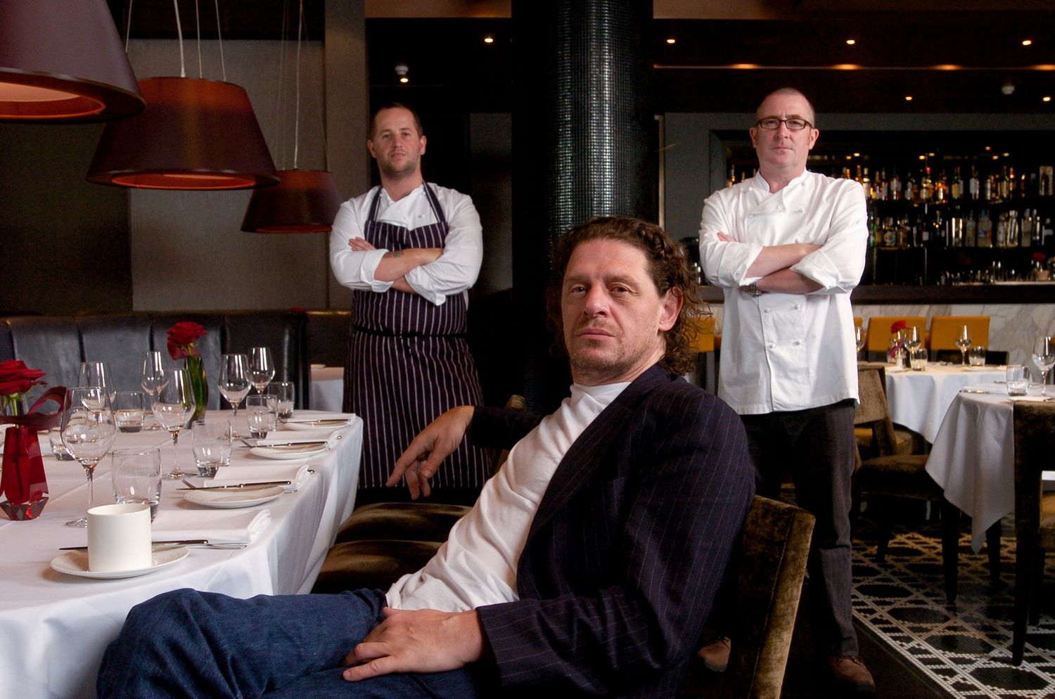 Three men pose in an elegant restaurant, two wearing chef uniforms and one in casual attire near a set dining table.