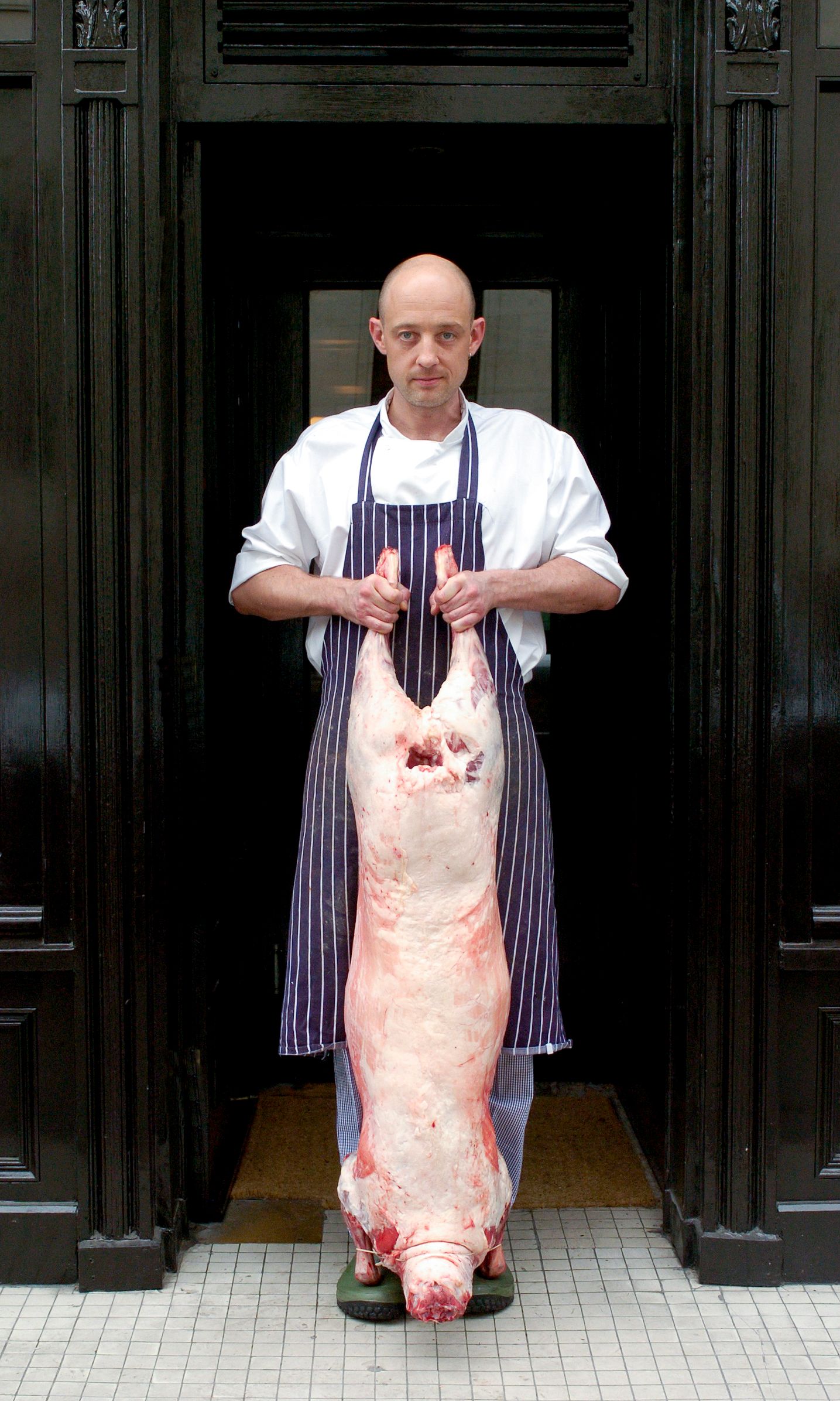 A bald chef in a white coat and striped apron holds a whole raw pig carcass while standing in a doorway.