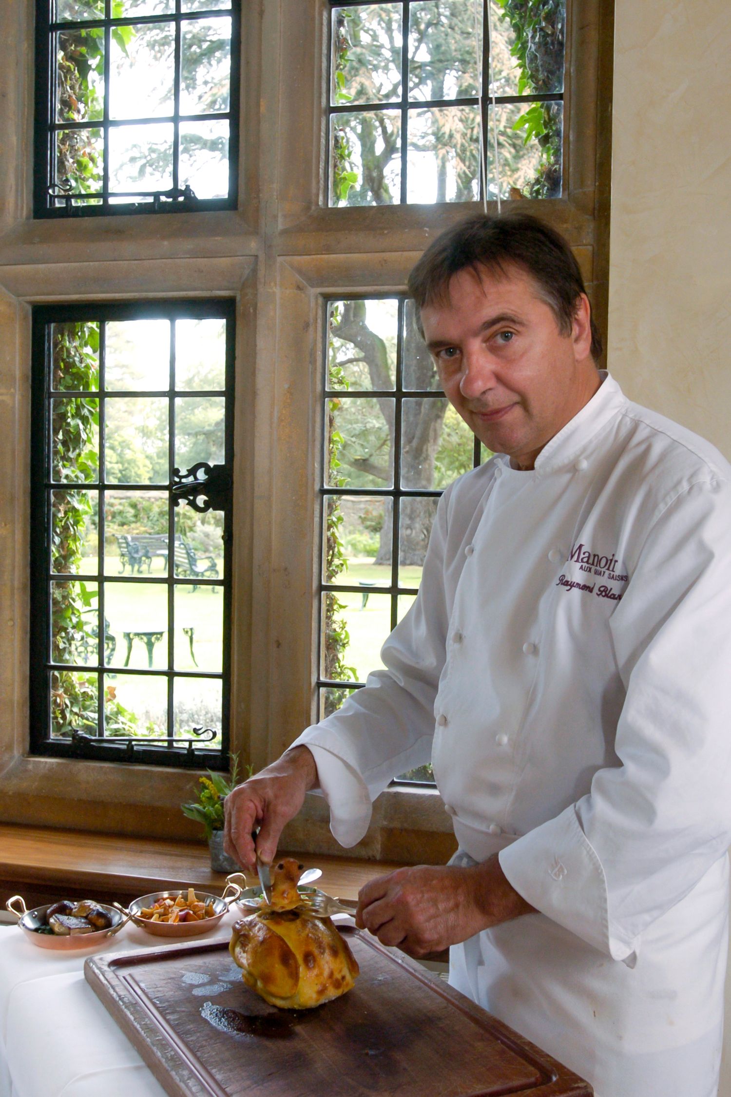 Chef in white uniform plating gourmet dish on wooden board near leaded glass windows with garden view.