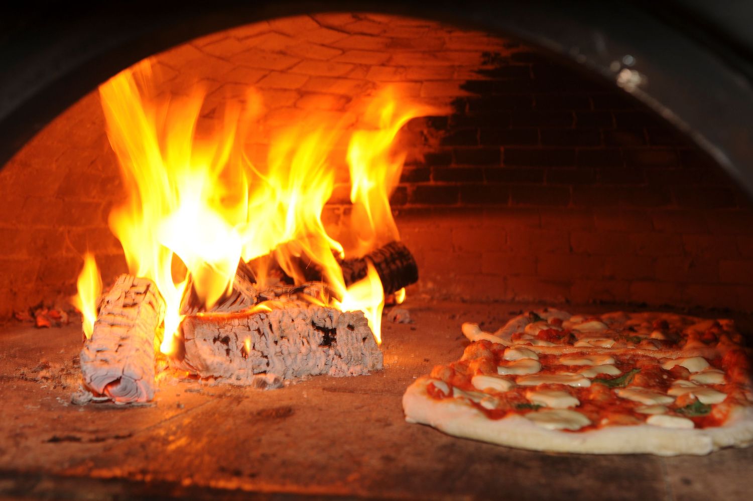 Pizza baking in a traditional wood-fired oven with open flames and glowing embers.