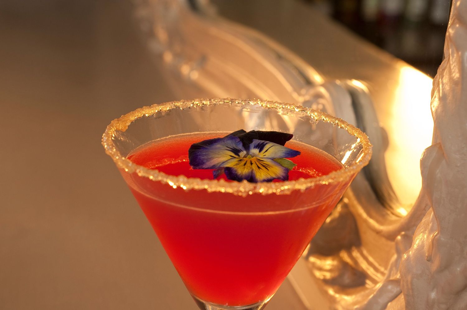Red cocktail in a sugar-rimmed martini glass garnished with a purple pansy flower, served on a silver tray.
