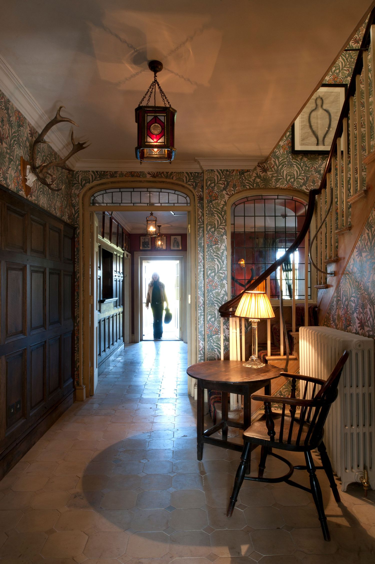 Historic hallway with ornate wallpaper, antler decor, hanging lantern, Windsor chair, and lamp on round table.