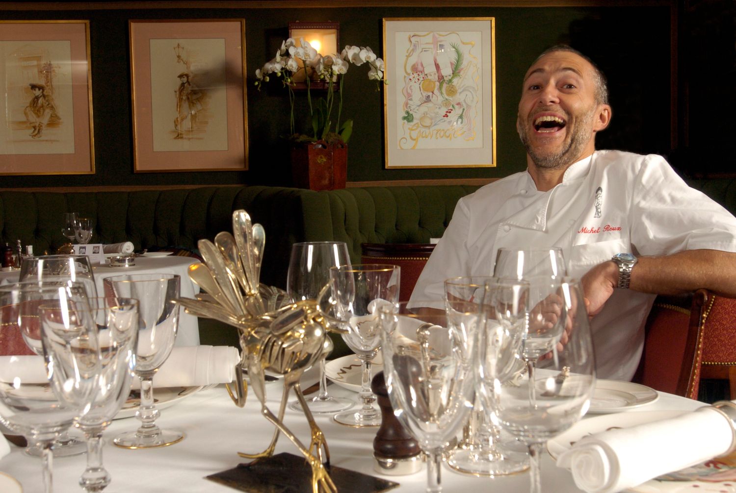 Chef in white uniform laughing joyfully at elegantly set restaurant table with wine glasses and golden cutlery.
