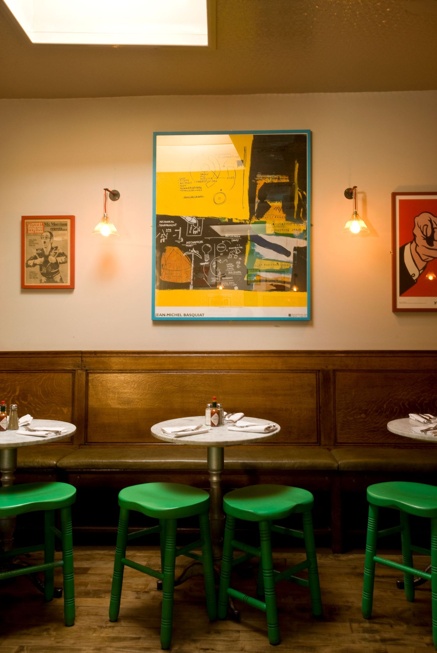 Cozy restaurant interior with green stools, white tables, wood paneling, and colorful modern art prints on beige walls.