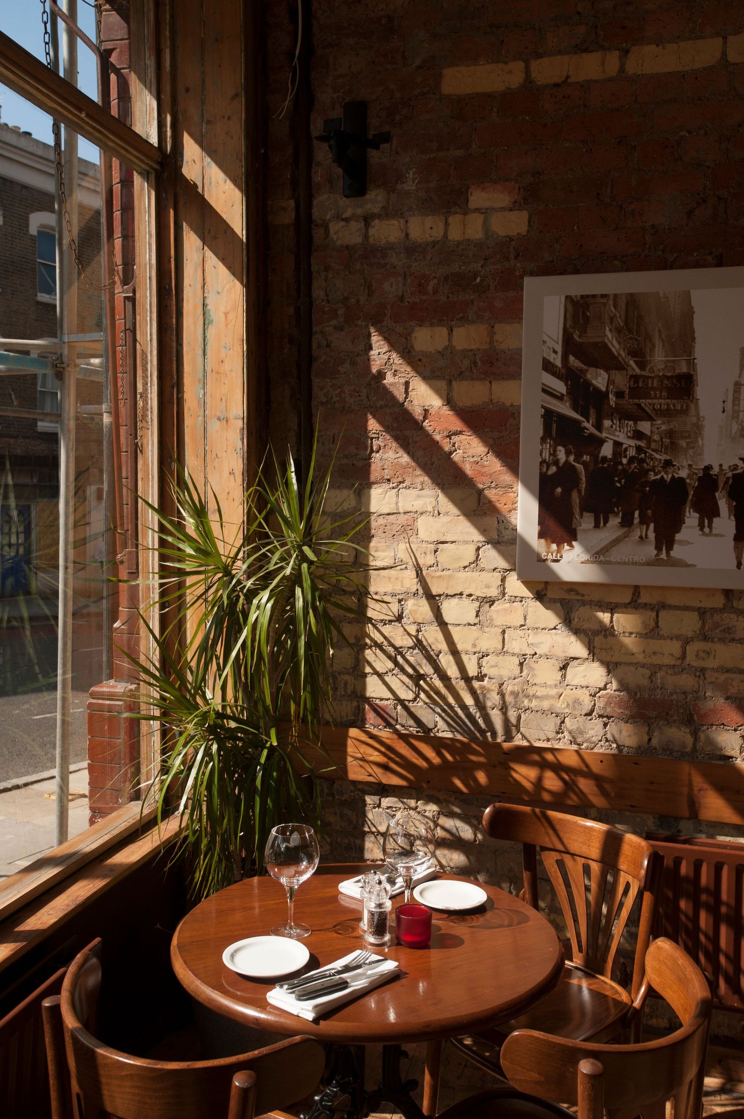 Sunlit rustic restaurant corner with brick walls, round wooden table set for two, wine glasses, and a tropical plant by large windows.