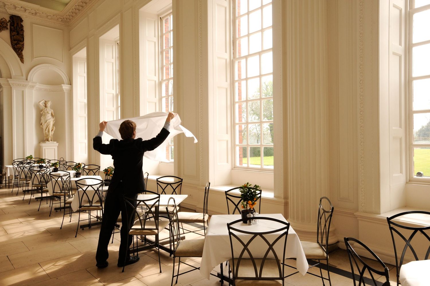 A waiter shakes out white napkins in an elegant sunlit dining room with tall windows and neatly arranged tables and chairs.