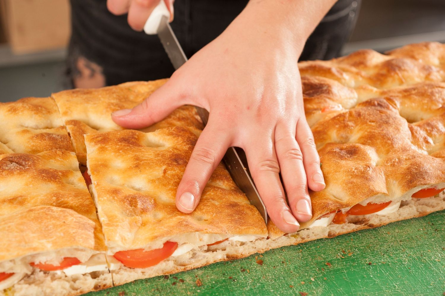 A person slicing a freshly baked flatbread pizza with tomatoes into portions on a green surface.