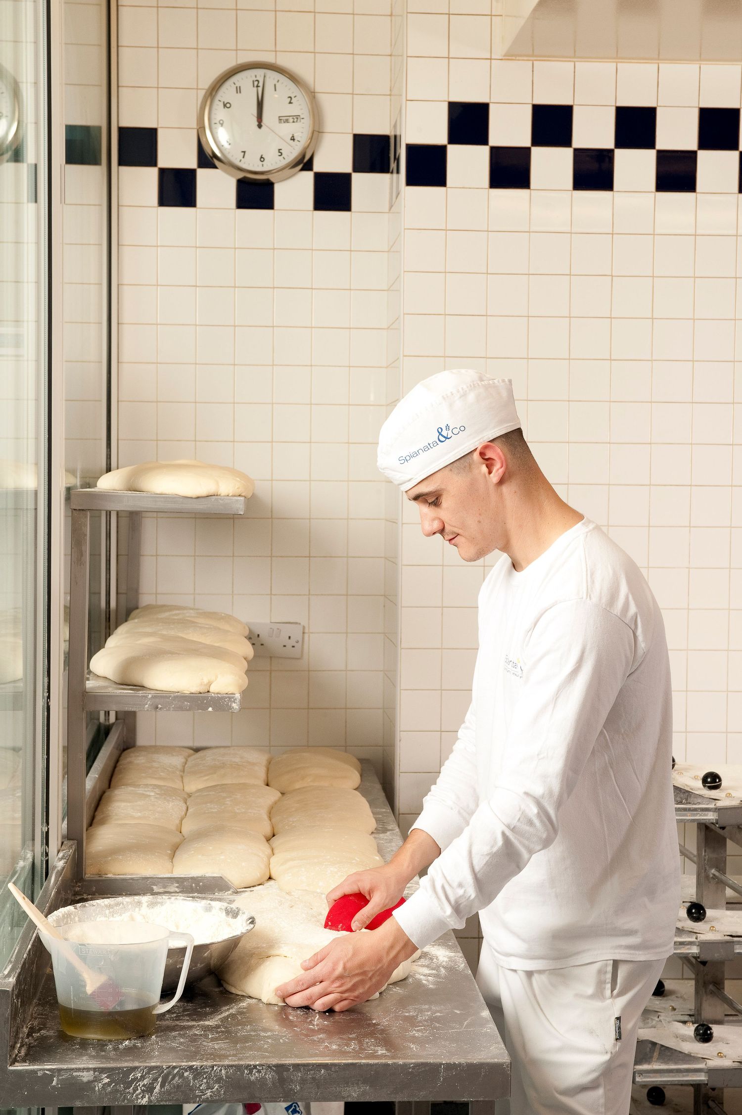 A baker in white uniform shapes bread dough on a metal counter in a tiled bakery kitchen with a clock on the wall.