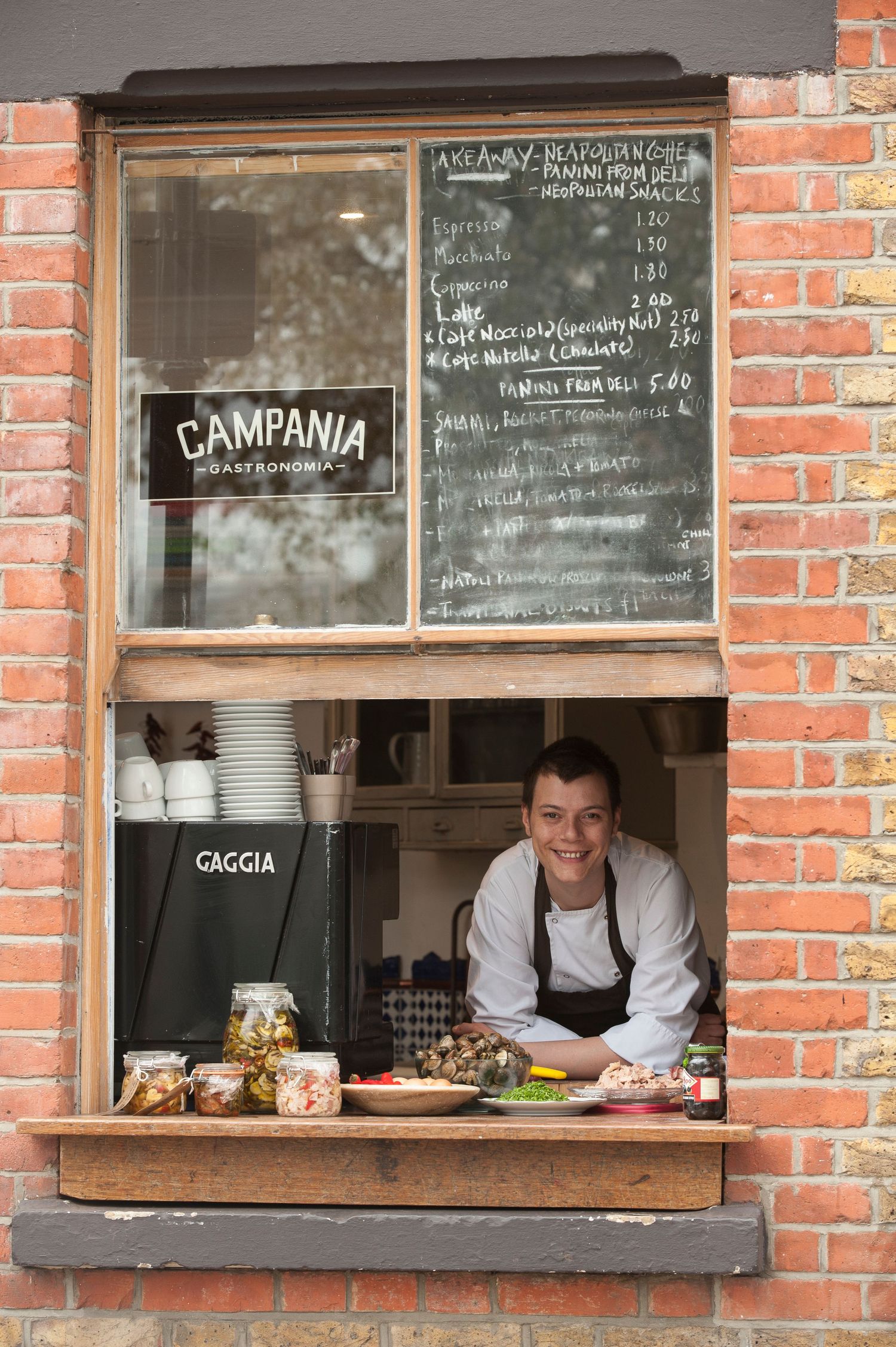 Chalkboard menu and service window in brick wall with restaurant staff visible inside.
Photograph by Danny Elwes