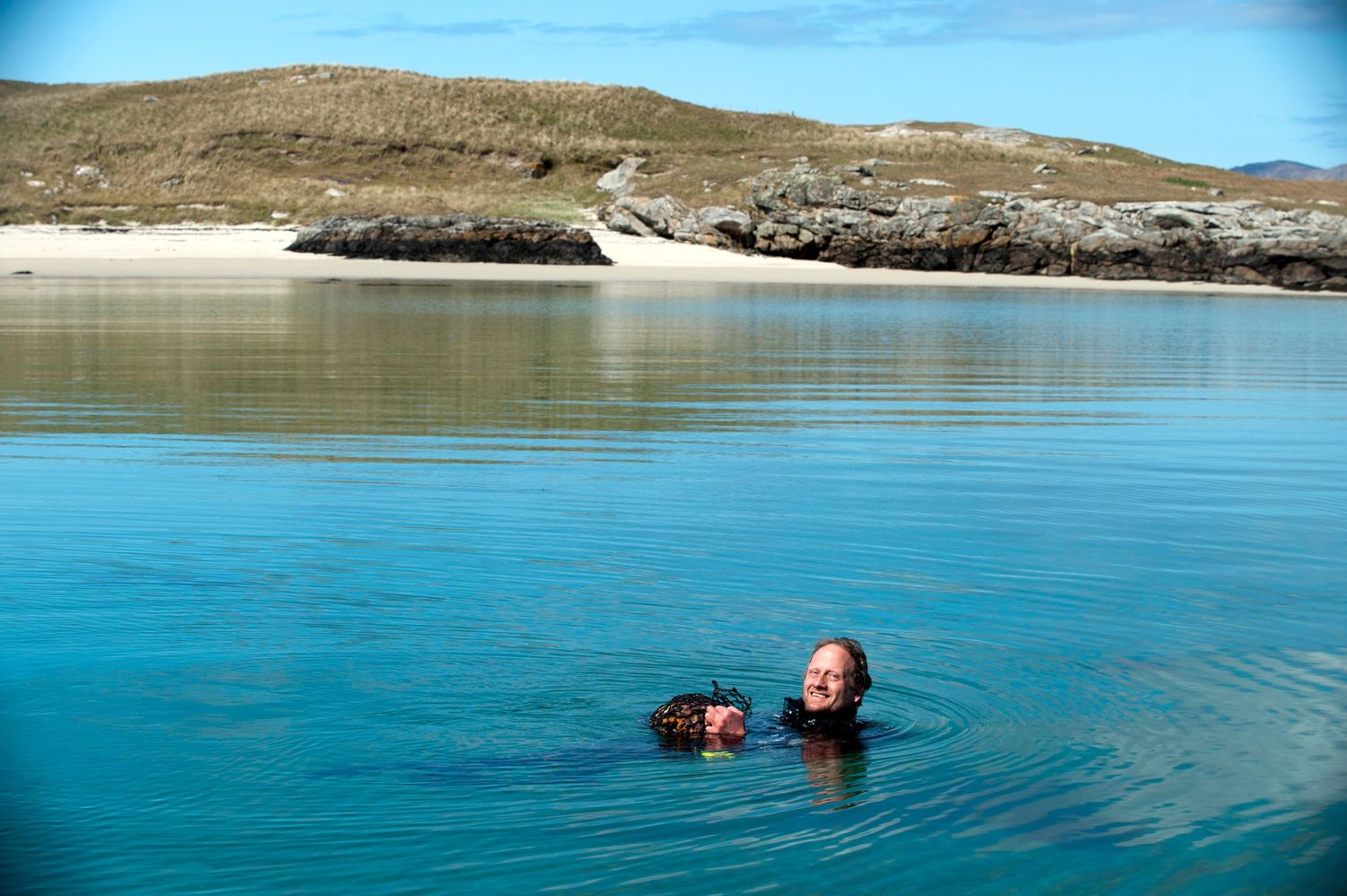 Two people swimming in clear turquoise water near a sandy beach with rocky hills in the background.