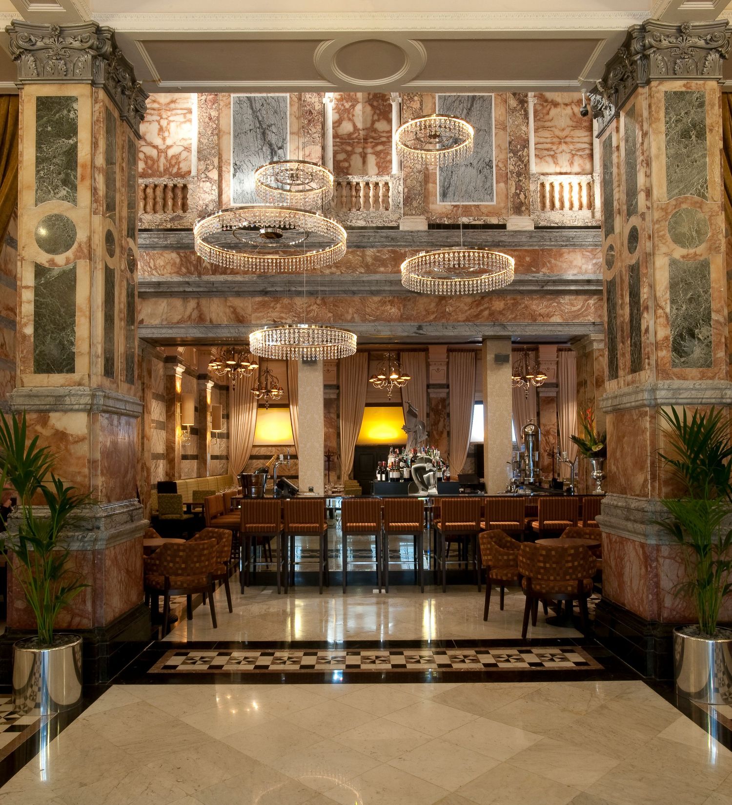 Elegant hotel lobby with crystal chandeliers, marble columns, checkered floors, and ornate decor in a grand classical style.