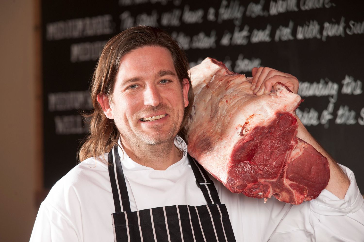 Smiling chef in apron holding a large cut of raw beef on his shoulder in front of a chalkboard menu.