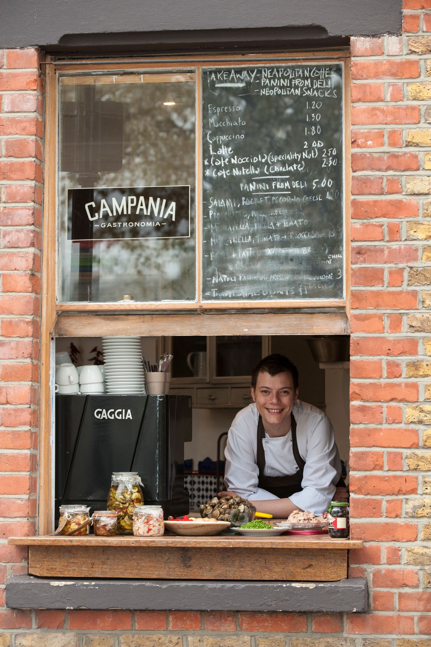 Smiling chef at Campania Gastronomia street food window with chalkboard menu on brick wall.