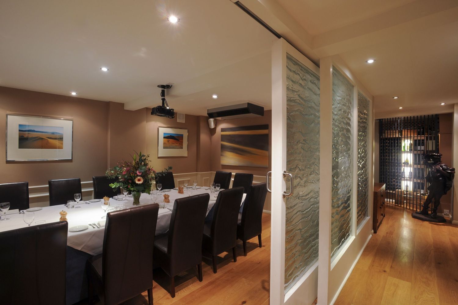 Elegant private dining room with long table, dark chairs, glass partition doors, hardwood floors, and warm recessed lighting.