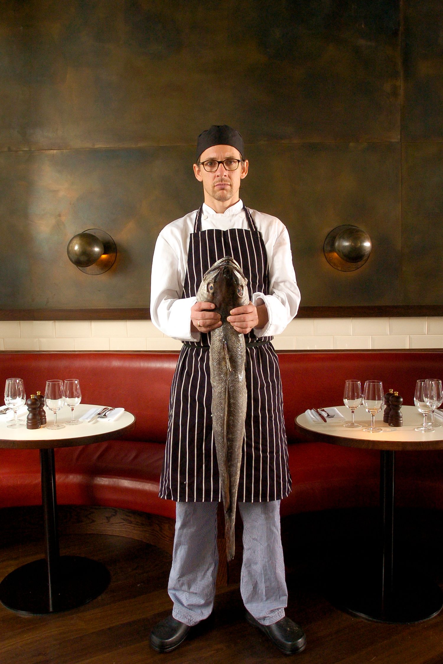 Chef in striped apron and glasses holds fresh whole fish in elegant restaurant dining room.