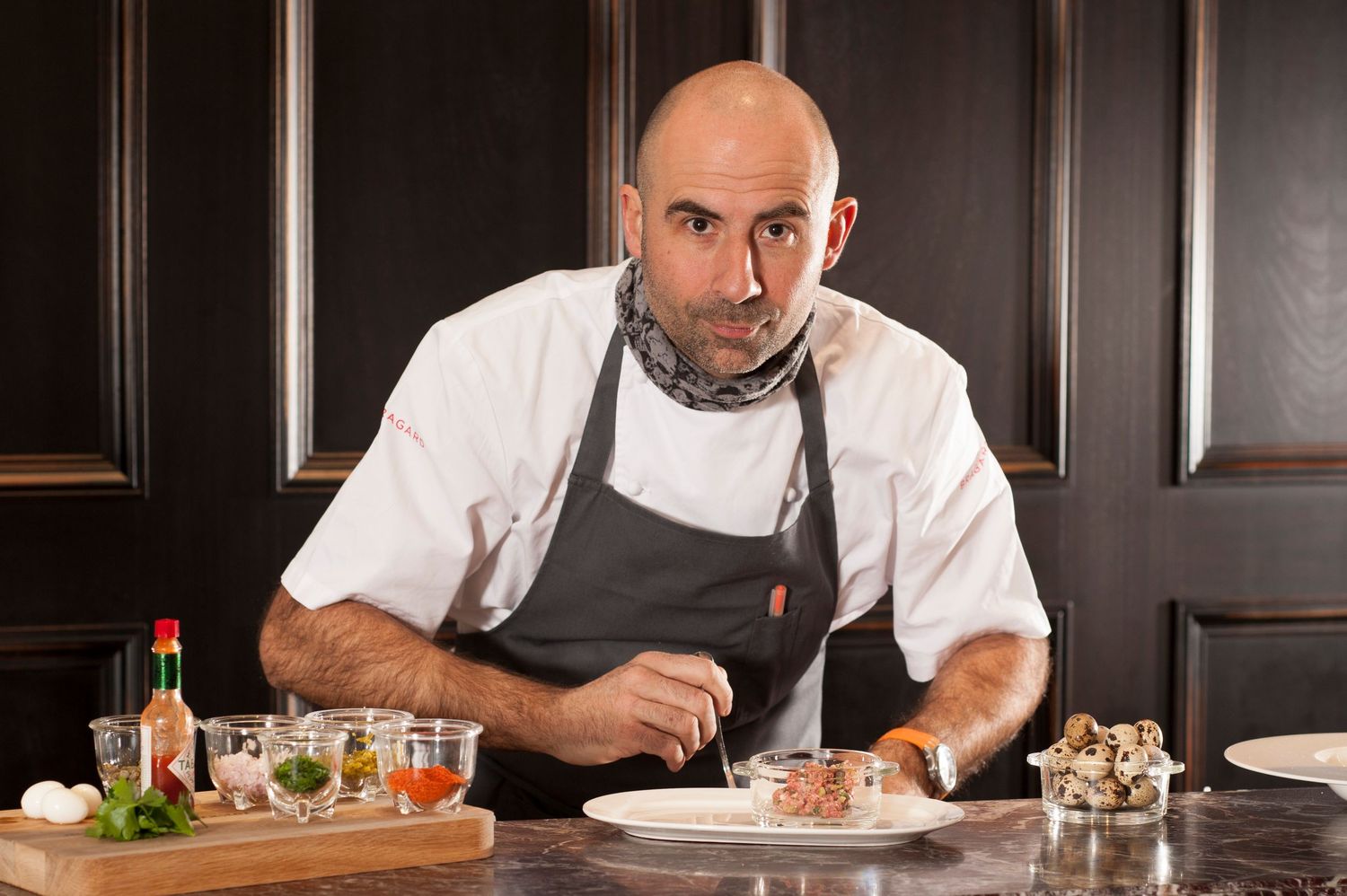 Bald chef in white coat and gray apron leans over a plated dish at a dark wood restaurant table with ingredients nearby.