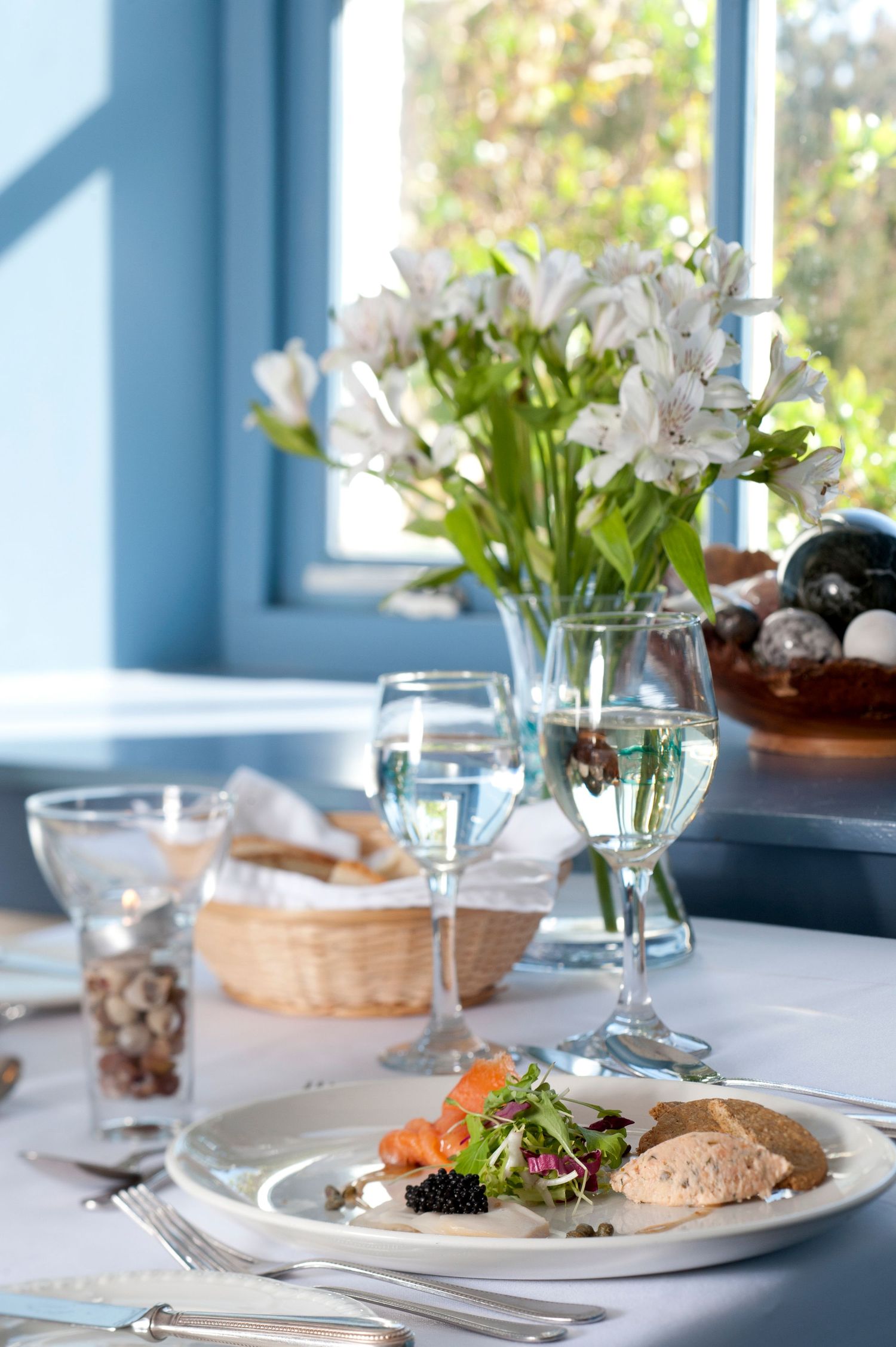 Elegant restaurant table setting with white alstroemeria flowers, wine glasses, and a gourmet salad plate by a blue window.