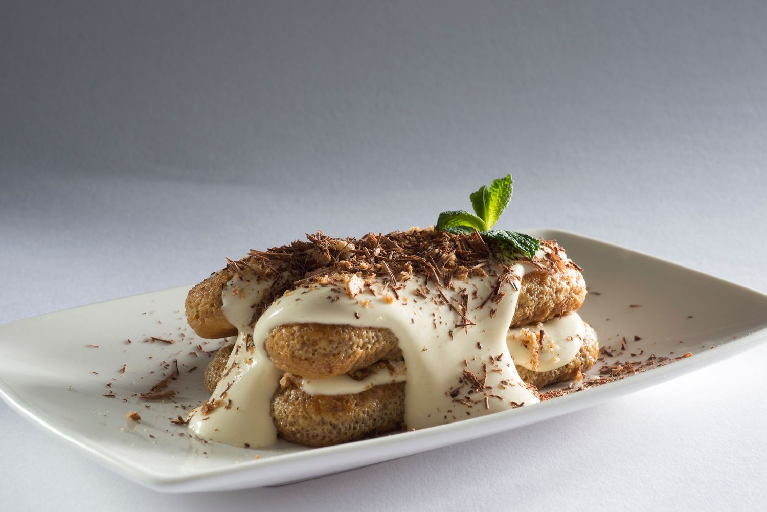 Classic tiramisu dessert with ladyfingers, mascarpone cream, chocolate shavings, and fresh mint on a white plate.