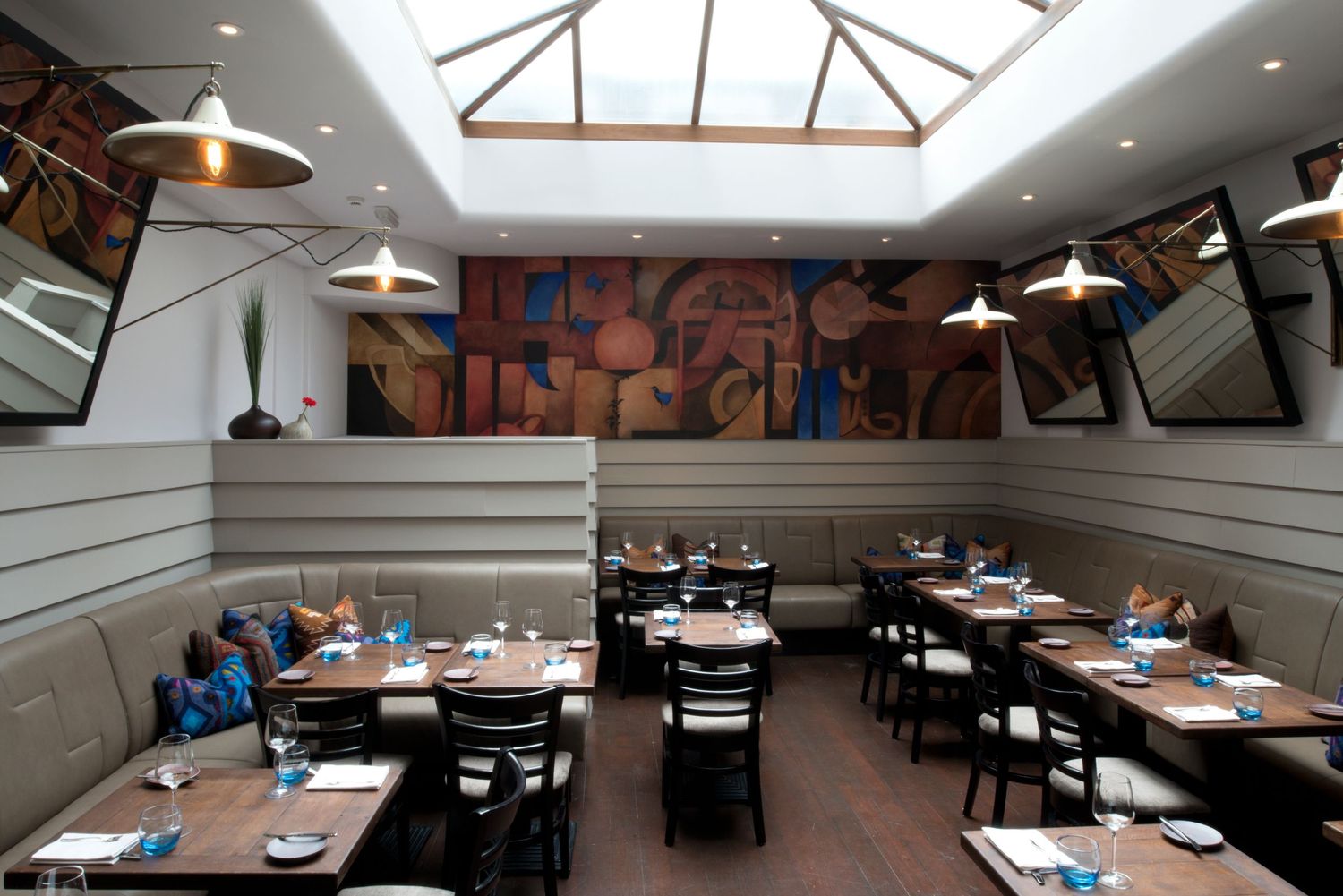 Elegant restaurant interior with wooden tables, pendant lights, skylight ceiling, and a colorful mural on the wall.