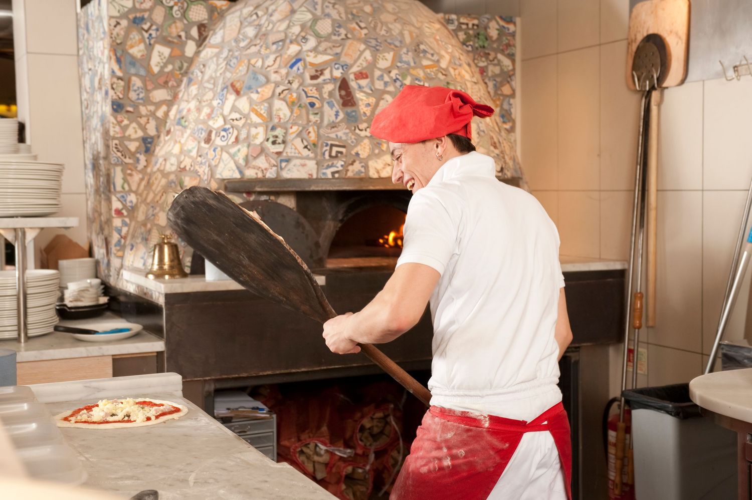 Pizza chef in red cap using wooden peel to place pizza in traditional wood-fired oven in restaurant kitchen.