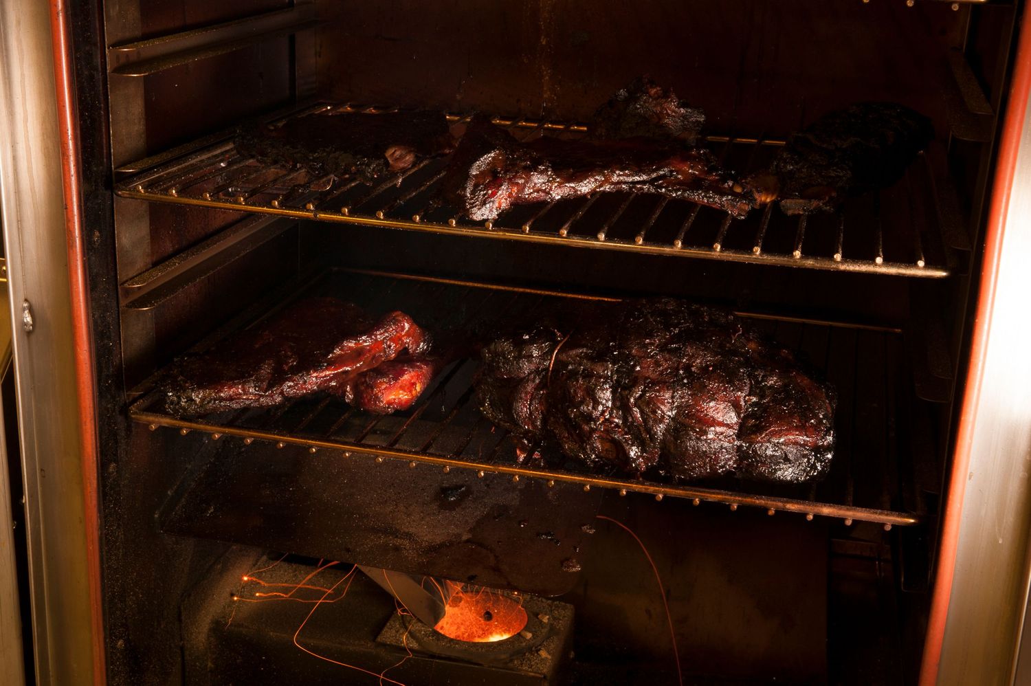 Smoky BBQ ribs and meat slow-cooking on racks inside a dark commercial smoker oven.