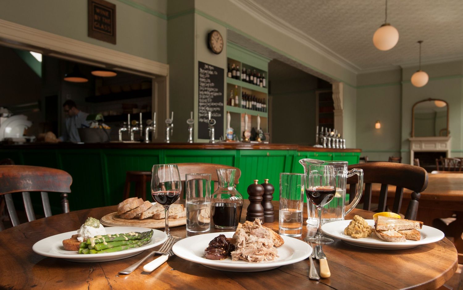 Elegant pub dining table set with plates of food, wine glasses, and water jugs before a green bar backdrop.