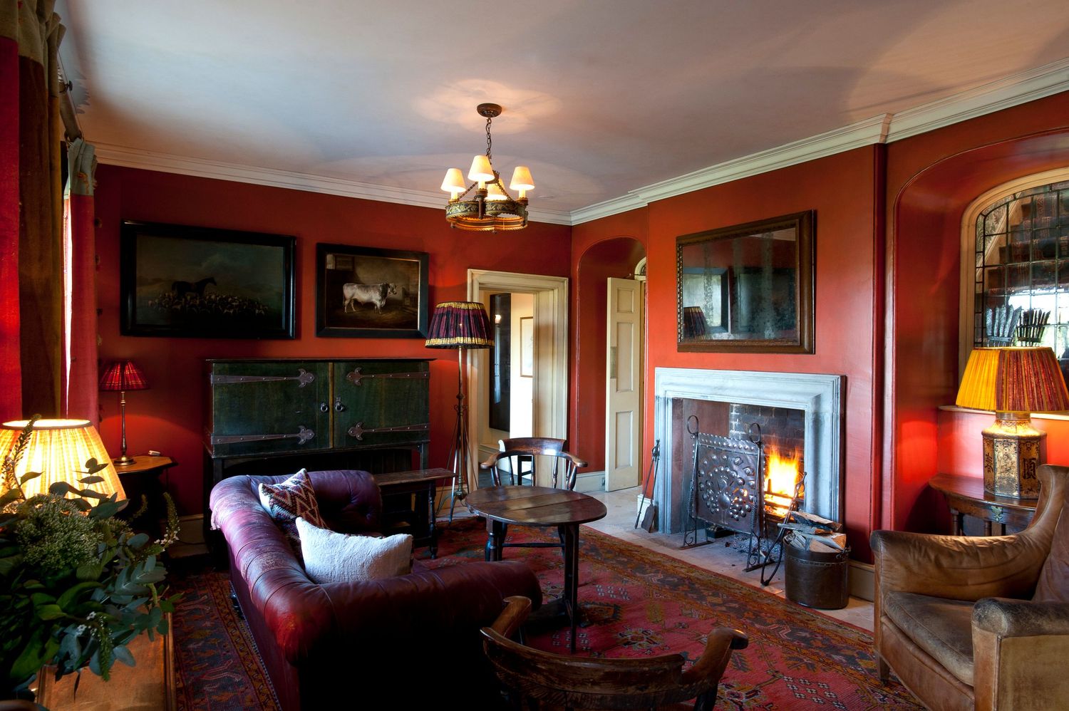 Elegant red-walled sitting room with lit fireplace, chandelier, leather sofa, Persian rug, and oil paintings in a classic interior.