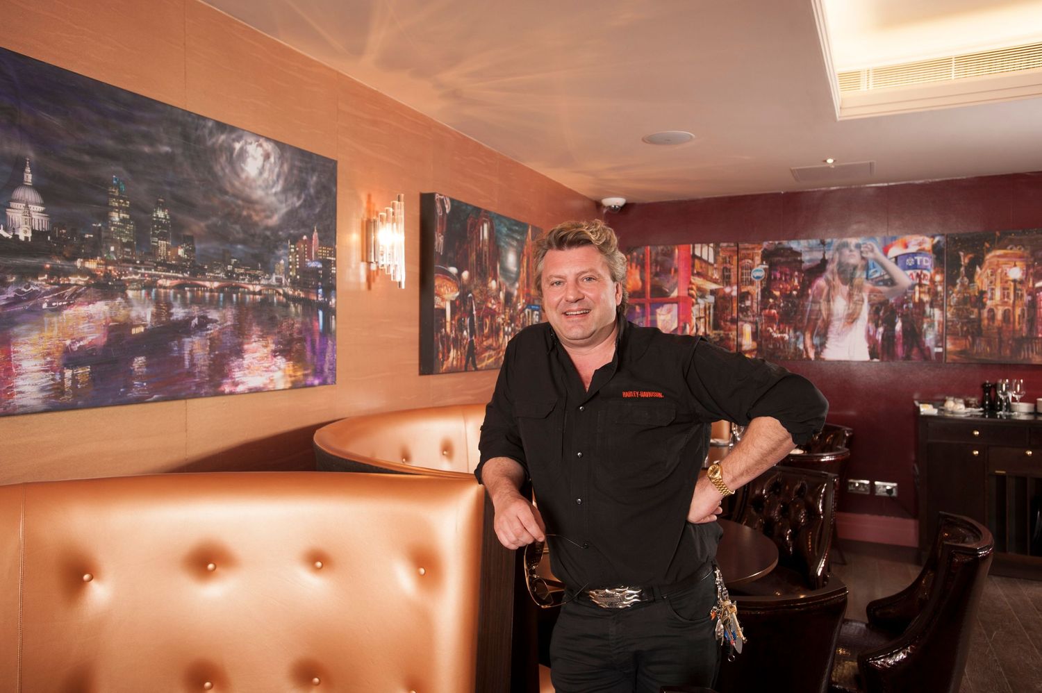 Man in black shirt posing in upscale restaurant with colorful city nightscape paintings on warm golden walls.