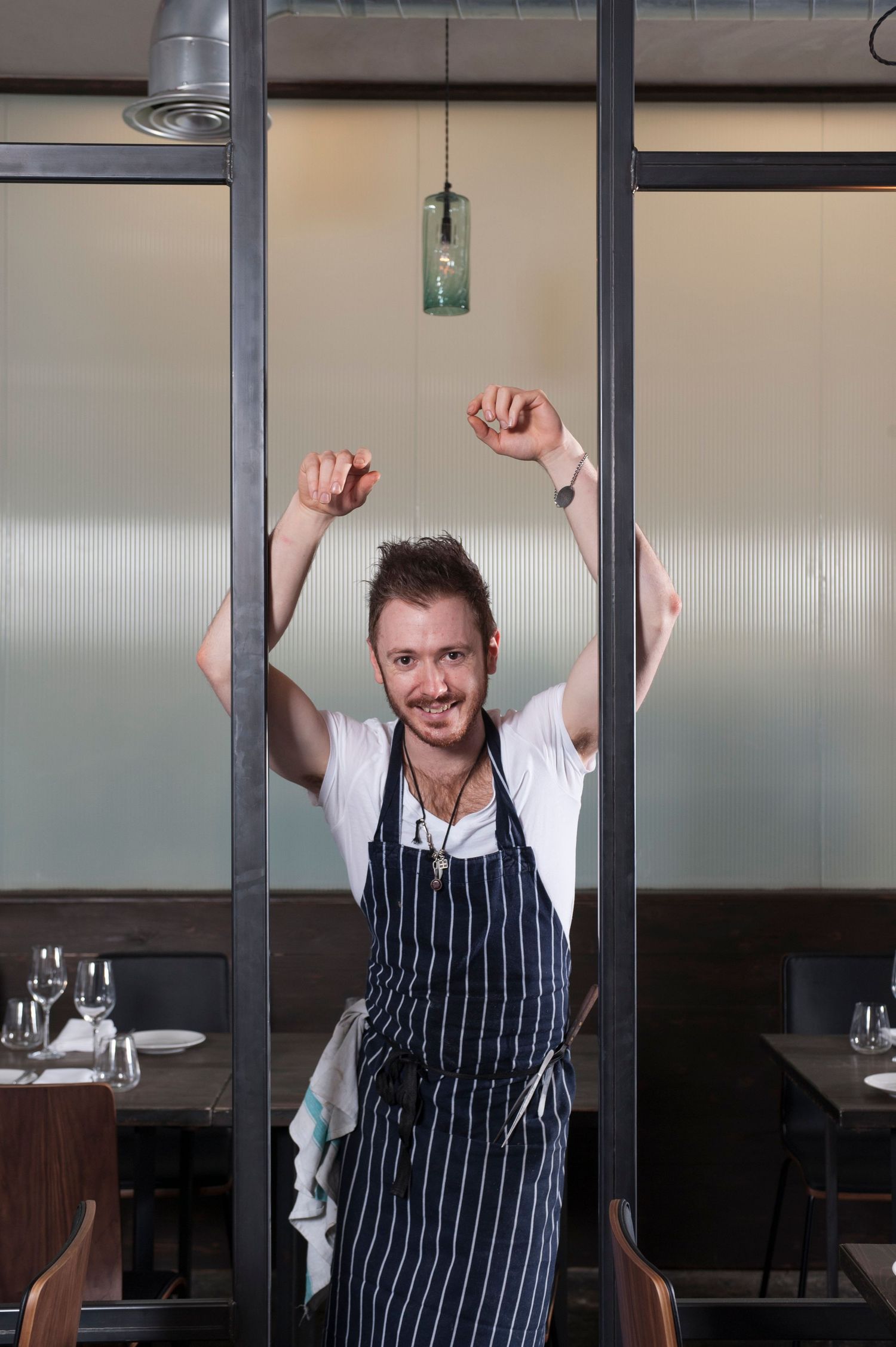 A cheerful chef in a striped apron raises his arms joyfully inside a modern restaurant dining room.