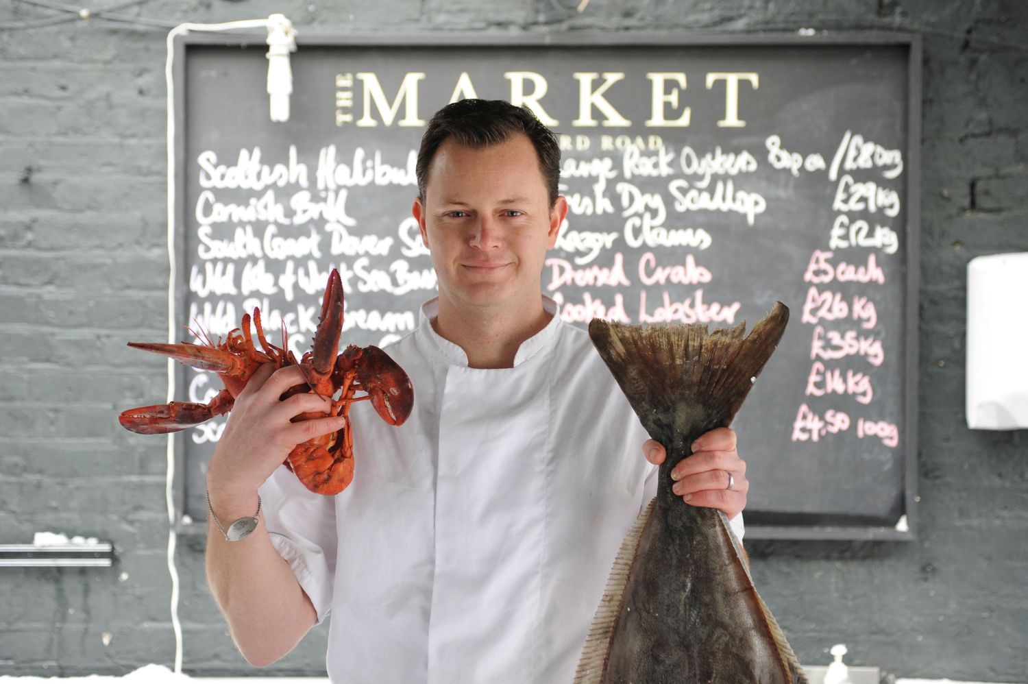 Chef in white coat holds fresh lobster and halibut in front of market chalkboard menu.