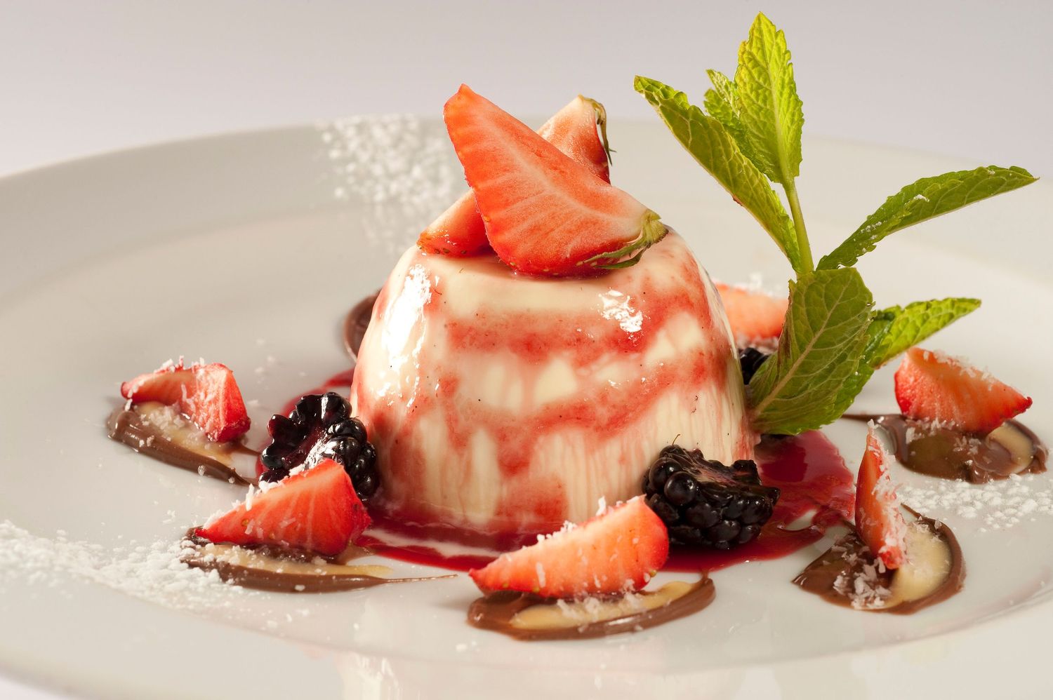 Elegant panna cotta dessert topped with fresh strawberries, blackberries, red berry sauce, and mint on a white plate.