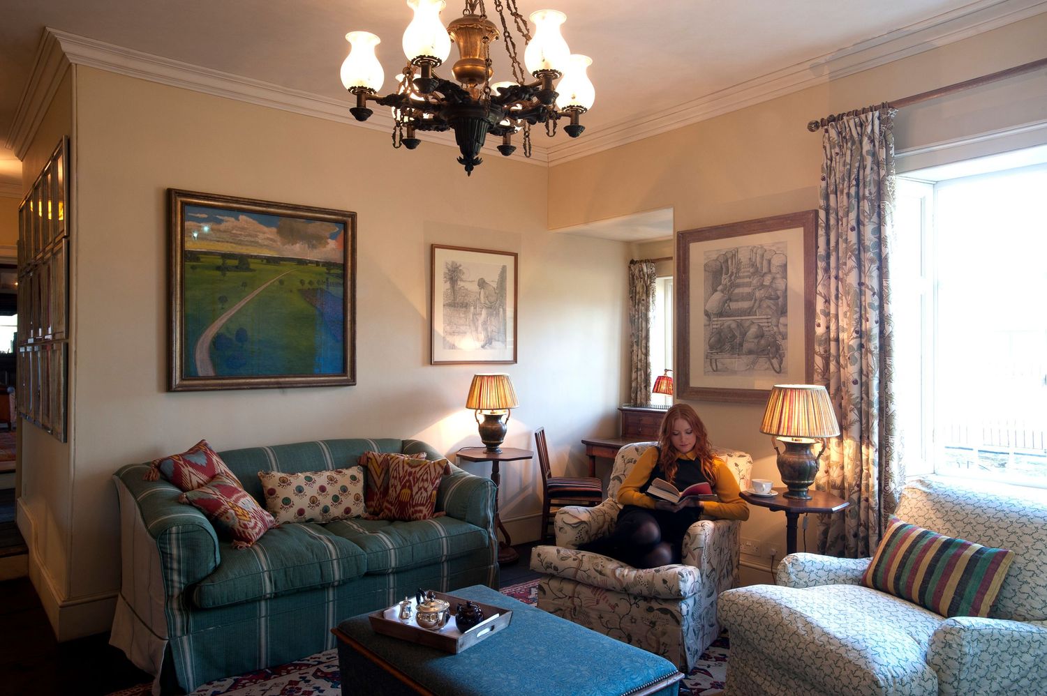 Elegant living room with teal sofas, floral armchairs, chandelier, and a woman reading near a bright window.