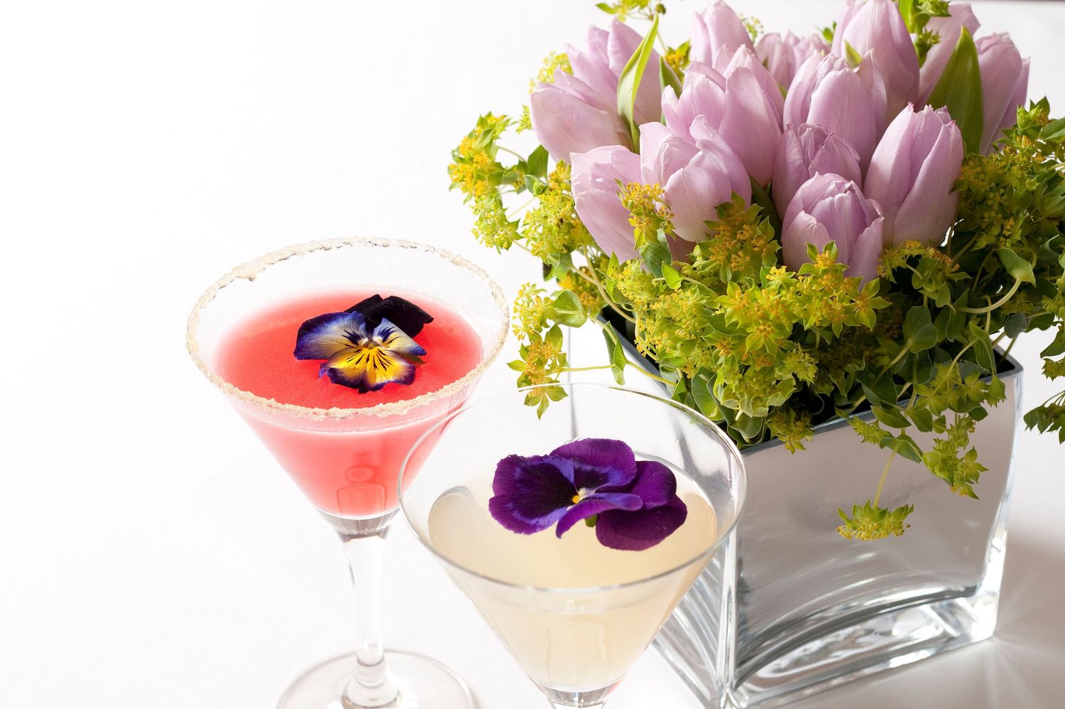 Two elegant cocktails garnished with edible pansies beside a vase of pink tulips and yellow flowers on white background.