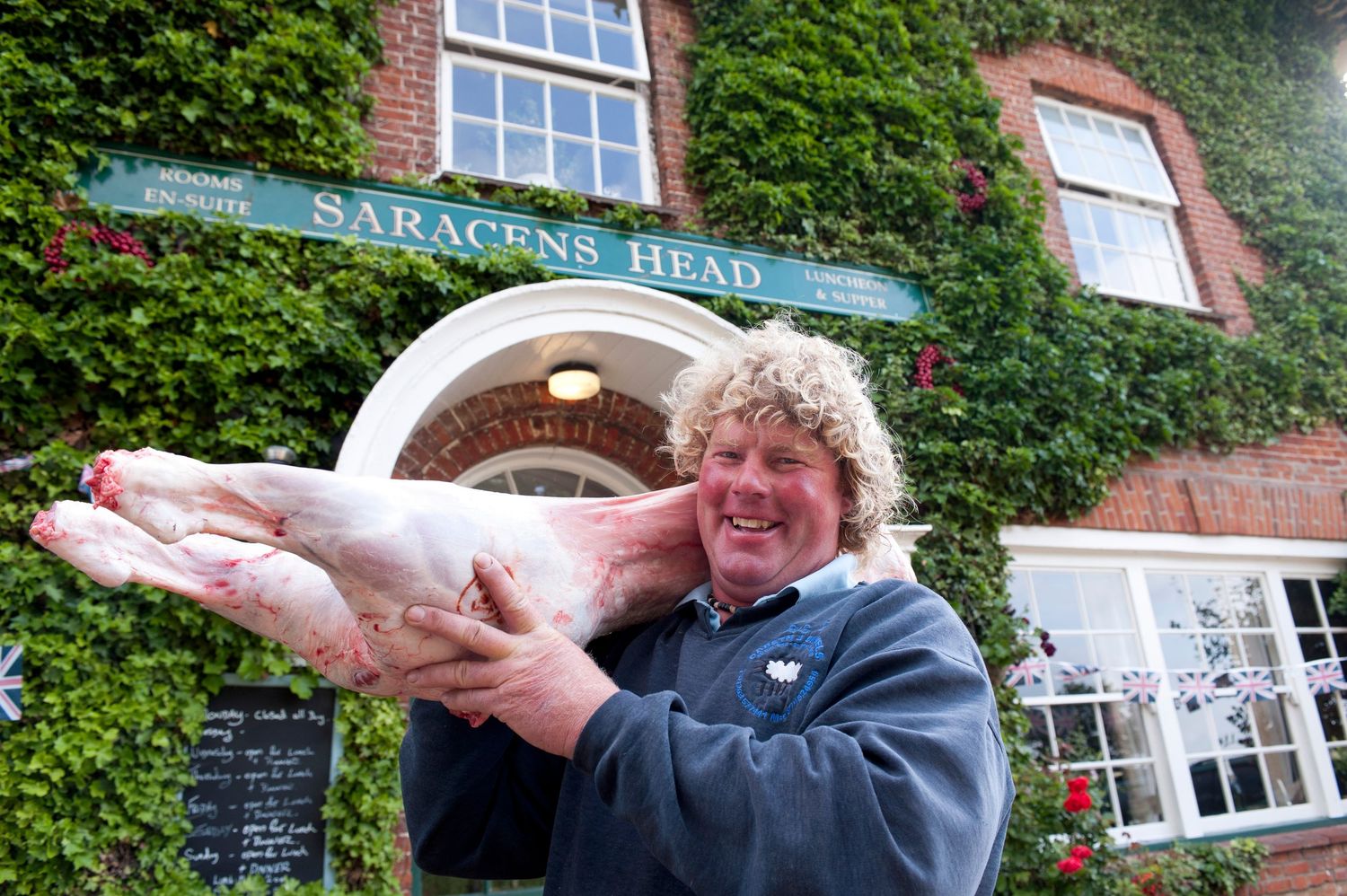 Man holding a whole raw pig outside the Saracens Head pub, smiling proudly at the camera.