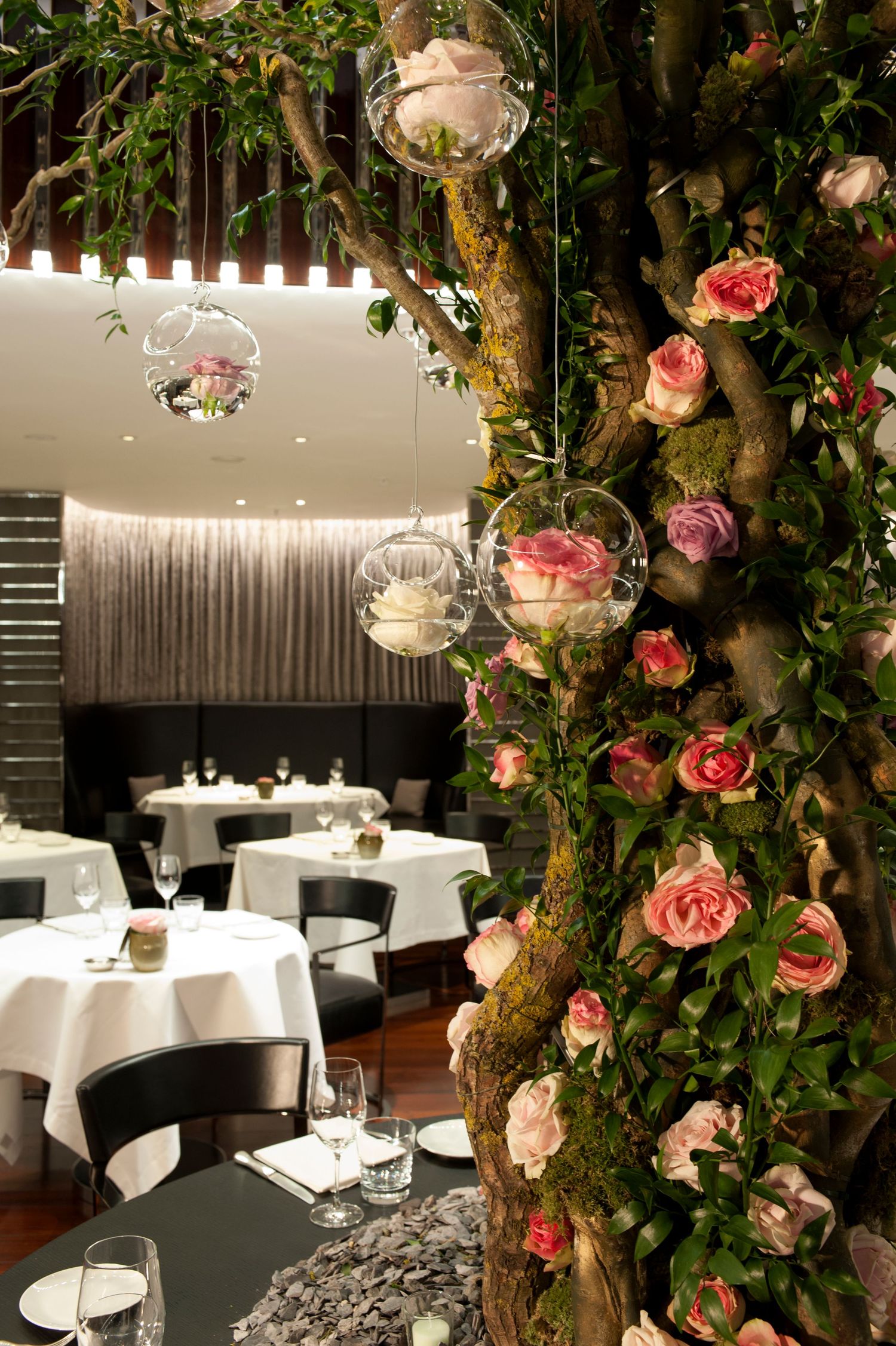 Elegant restaurant interior with rose-covered tree, hanging glass globe terrariums, and white-clothed dining tables in soft lighting.