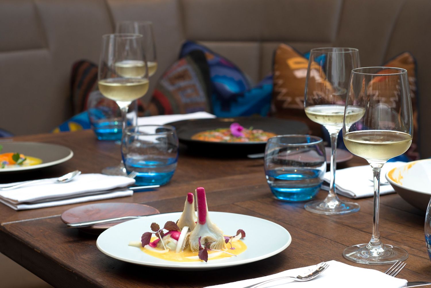 Elegant fine dining table set with white wine glasses, gourmet plated dishes, and blue water glasses on a wooden table.