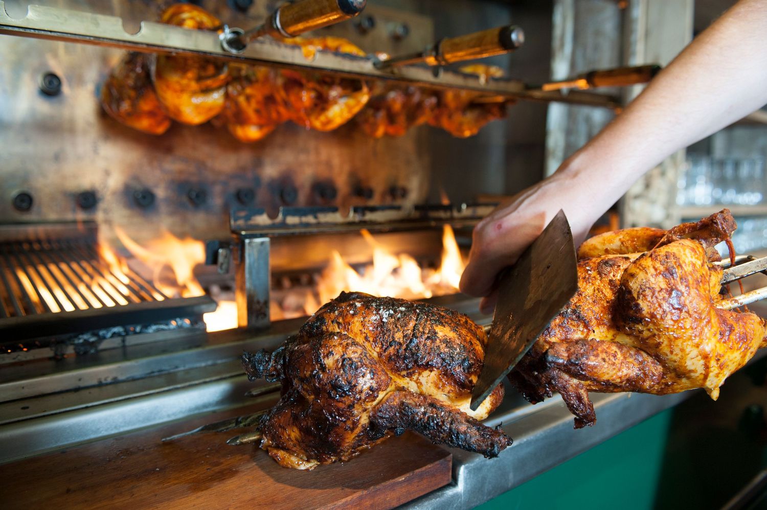 Rotisserie chickens roasting over open flames on a spit, with a hand using tongs to handle golden-brown grilled chicken.