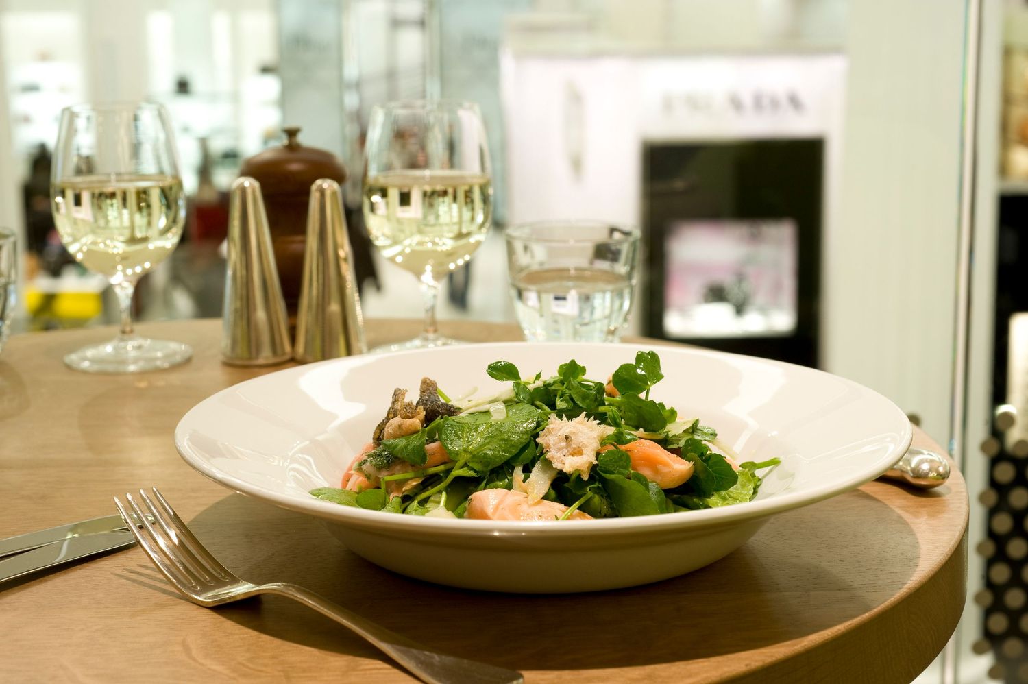 Fresh shrimp and greens salad served in a white bowl beside two glasses of white wine at an elegant restaurant.