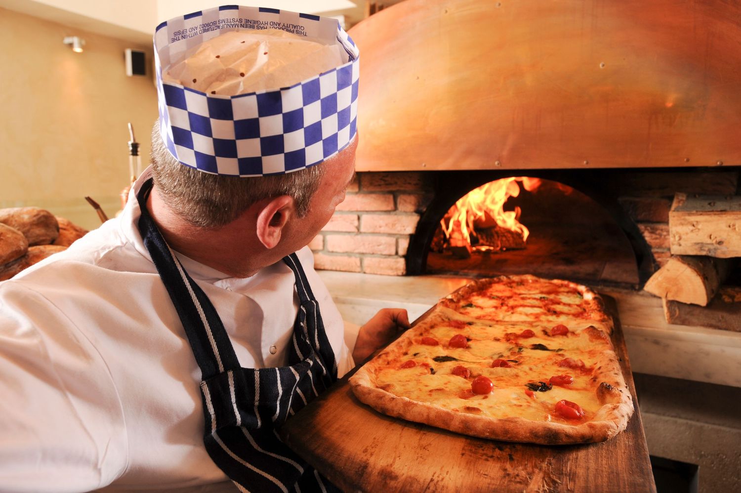 Chef in checkered hat slides a fresh Margherita pizza into a wood-fired brick oven using a wooden peel.