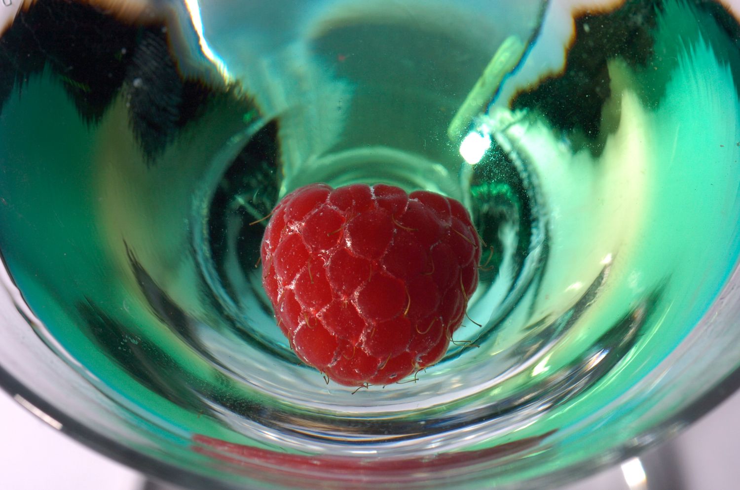 A fresh red raspberry sits at the bottom of a green cocktail in a clear martini glass.