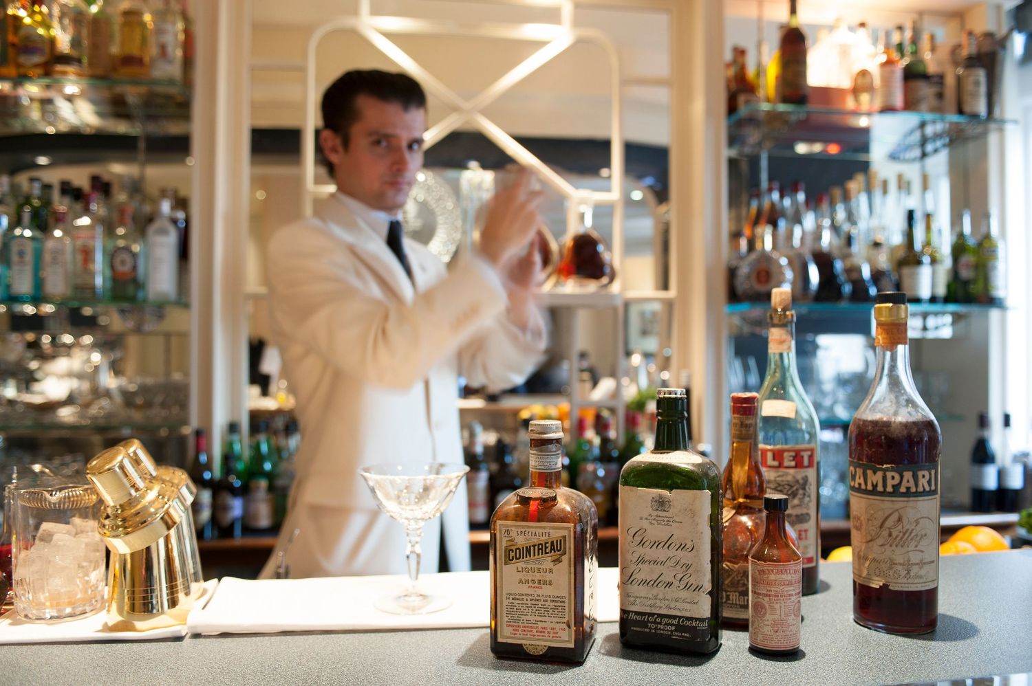 Bartender in white jacket shaking cocktail behind bar with Campari and vintage spirit bottles on counter.
