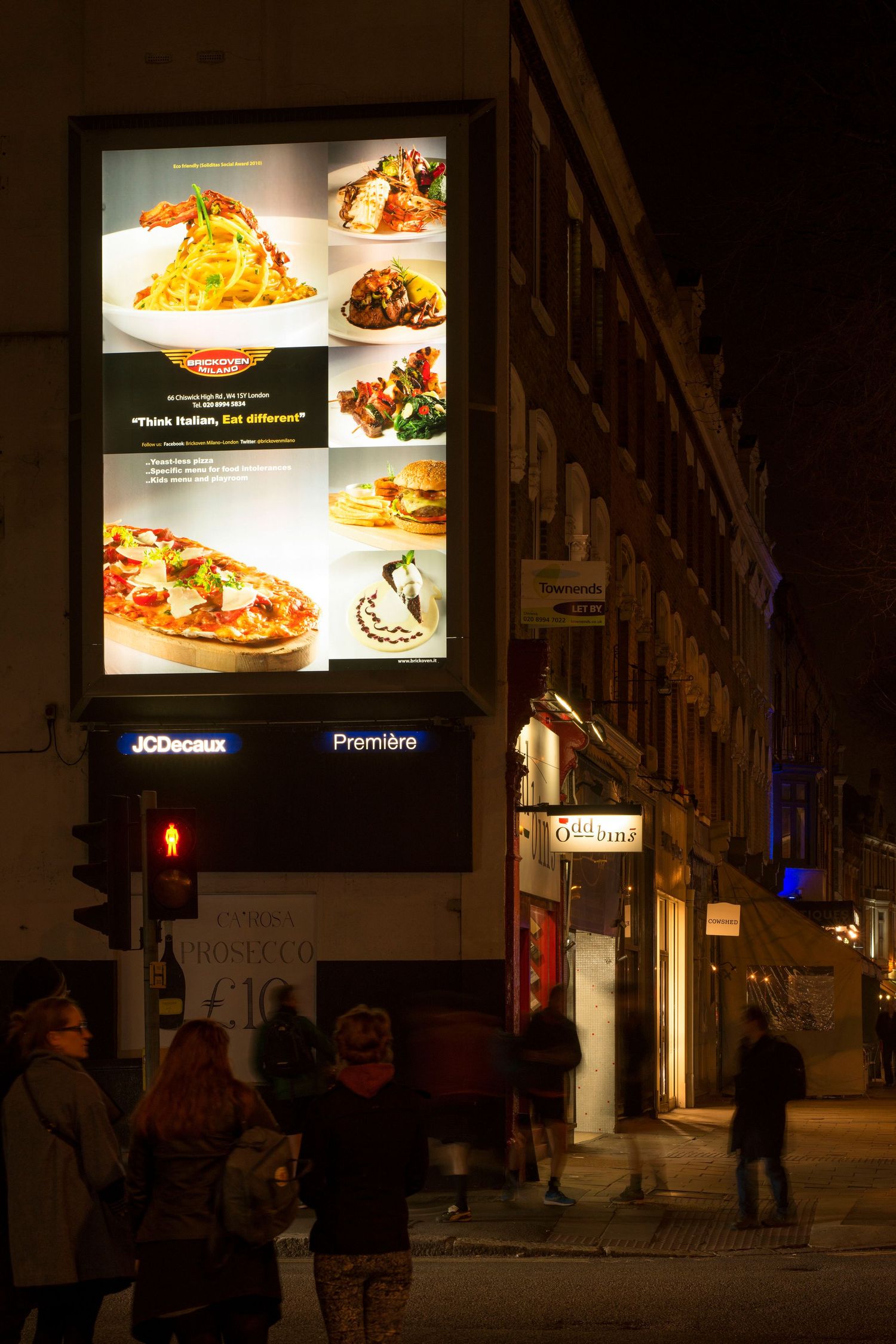 Bright outdoor JCDecaux food menu billboard displaying Italian dishes at night with people gathered on the street.
